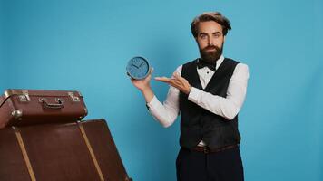 Classy bellhop poses with wall clock, looking at time and panicking for running late in tourism industry. Punctual hotel concierge employee using watch to see exact hour and minutes. photo