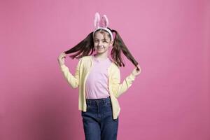 Cute small girl posing in studio with bunny ears and pigtails, celebrating easter and spring time. Adorable cheerful child standing over pink background, seasonal traditions and festive toys. photo