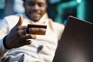 Close up shot of debit card in man hand adding payment method on website. Focus on credit card held by person in blurry background doing online shopping from neon lit apartment photo