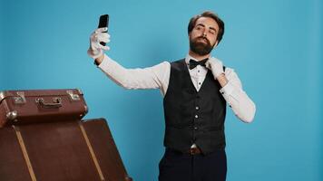 en estudio, portero en traje toma fotos mientras utilizando teléfono inteligente aplicación para selfies y interino tonto en contra azul antecedentes. elegante profesional hotel conserje haciendo imágenes con su teléfono.