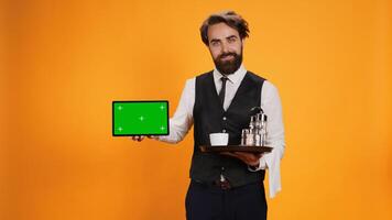 Joyful employee shows greenscreen on tablet, posing with food platter against yellow background. Five star waiter presenting blank isolated mockup template on display in studio, formal attire. photo