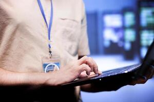 Worker in front of blurry background supercomputers delivering web content to online customers. Repairman in server room using laptop to make sure critical systems are working errorless, close up photo