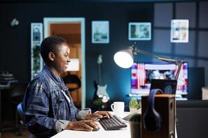 sonriente negro mujer gerente un complejo sistema utilizando máquina aprendizaje en su personal computadora. alegre africano americano programador sentado y mecanografía en código para creando base de datos en ordenador portátil. foto