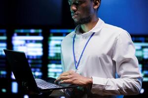 African american IT developer using laptop to examine server mainframes providing vast computing resources. Data center enabling AI to process massive datasets for complex machine learning operations photo