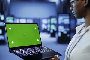 Programmer in server room uses green screen laptop to future proof network from downtimes and unexpected system failures. Tech support with mockup device ensures increased data security photo
