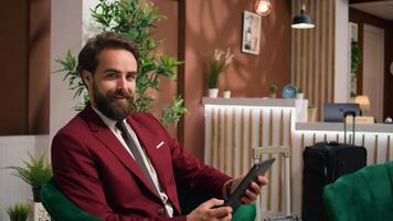 Entrepreneur in hotel lounge area using tablet to review new financial notes before attending official shareholders meeting. Young adult waiting to check in at resort, wearing tailored suit. photo