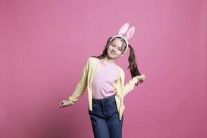 Cheery little girl dancing and having fun in front of camera, feeling happy and excited about easter celebration. Confident small toddler showing dance moves while she wears bunny ears. photo