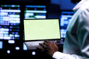 Computer scientist using green screen laptop to verify configuration settings of data center hardware. Repairman utilizes monitoring software to troubleshoot root cause of defects in server mainframes photo