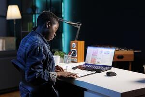 Dedicated African American lady analyzes financial data on her laptop and paperwork. Black woman carefully studies market trends and makes profitable investment decisions with precision and focus. photo