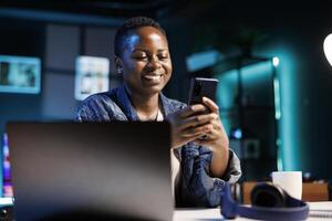 Cheerful female college student using mobile device to engage with friends on social media platforms. African american lady using cell phone to surf the web while sitting at desk in her home office. photo