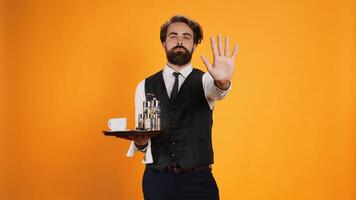 Skilled butler indicates stop sign with palm, posing against yellow background with food tray. Restaurant worker in suit showcasing rejection and dismissal, disagreeing with something. photo