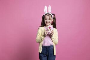 Cute young kid posing with a pink stuffed rabbit toy in studio, presenting her decorations in time for easter celebration. Small girl with pigtails holding a colorful bunny on camera. photo