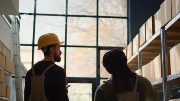 Meticulous trainee walking through warehouse rows scanning cardboard boxes on shelves under team leader guidance, using digital devices to update info before sending parcels for delivery photo