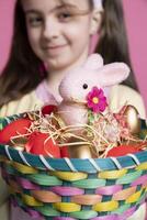 Young cheerful kid holding a colorful decorations basket on camera, showing painted eggs and a stuffed pink rabbit as festive arrangement. Cute girl smiling in studio with adorable decor. Close up. photo