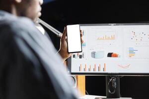 African american guy looks at white blank display while he works on financial operations with sales presentation. Male freelancer doing remote tasks from home, holding phone with empty layout screen. photo