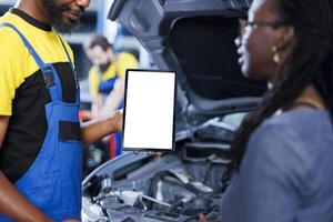 Repairman in car service uses mockup tablet to order new exhaust pipes for broken vehicle. African american garage expert shows client needed components replacement on isolated screen device, close up photo