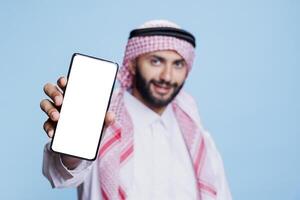 Muslim man wearing islamic headscarf and thobe smiling while holding smartphone with white screen in hand. Arab person showing mobile phone empty display for copy space closeup photo