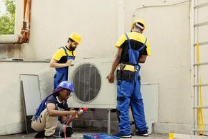 Diverse team of seasoned wiremen installing new external air conditioner for customer. Licensed servicemen tasked to optimize new HVAC system's performance, ensuring it runs at perfect efficiency photo