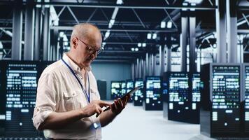 Senescent professional navigating network of server rigs in industrial nexus. Employee with tablet doing analysis and optimizations in high tech data center, preventing system perils photo