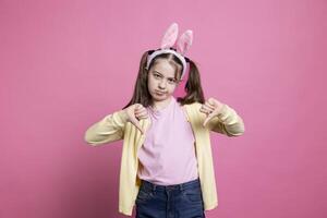 Negative displeased young kid showing thumbs down sign in studio, acting disappointed and sad before easter celebration. Mad angry small child being confident with her dislike symbol on camera. photo