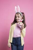 Adorable small kid holding a fluffy pink rabbit toy on camera, presenting her easter colorful toys and ornaments. Young happy child with bunny ears feeling joyful and excited about festivity. photo