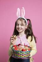 Small positive kid with bunny ears shows Easter toys and items, presenting exciting april festivities. Young cheerful girl with adorable pigtails showing a rabbit in a basket and colorful eggs. photo