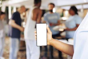 Close-up shot of african american person holding a mobile device showing blank copyspace mockup template. Detailed image of a smartphone with isolated white screen display at non-profit food drive. photo