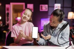 Happy girl next to her mother presents mockup smartphone in pink neon lit living room used as vlogging studio. Young media star helped by parent to showcase isolated screen phone photo