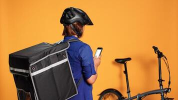 Courier waiting for order to be procesed before picking up food from restaurant for upcoming delivery. Woman checking phone app to find location of next address, isolated over orange studio background photo