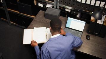 In evidence room, black police officer conducts an investigation using case files and personal computer. Male private detective examines witness accounts and documents to solve crime. Aerial shot. photo