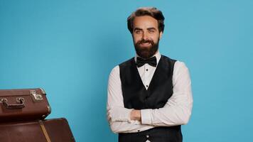 Professional hotel concierge posing on camera against blue background, preparing to carry luggage and help travellers. Elegant skilled employee working as bellboy, tourism industry concept. photo