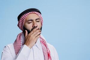 Exhausted muslim man wearing traditional headscarf and thobe yawning and covering mouth with hand. Tired person dressed in islamic ghutra headdress and robe making sleepy gesture photo