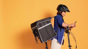 Handheld shot of BIPOC cyclist delivering takeout meal to clients, typing next address in phone app. Courier riding bicycle, wearing protective gear and thermal bag, isolated over studio background photo