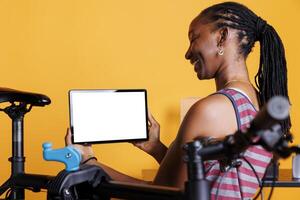 Sports-loving black woman stands by broken bicycle while carrying tablet with blank chromakey mockup template horizontally. African american female grasps digital gadget that displays a whitescreen. photo