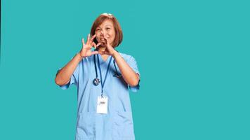 Portrait of cheerful healthcare professional doing heart symbol shape with hands. Licensed asian nurse showing love gesturing while at work, isolated over blue studio background photo