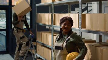 Portrait of smiling african american warehouse employee standing in front of shelves filled with packaged goods in cardboard boxes ready to be shipped, happy to work in professional establishment photo