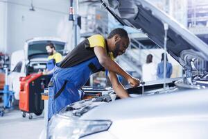 africano americano mecánico en coche Servicio usos esfuerzo de torsión llave inglesa a apretar nueces después reemplazando roto motor. proactivo garaje empleado usos profesional herramientas a reparar cliente automóvil foto