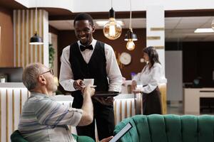 African american employee serving coffee to eldery male tourist with tablet in luxury hotel reception. Senior man holding a digital device and receiving a cup of tea from resort waiter in lounge area. photo