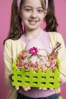 Positive excited small kid showing her handmade easter eggs in a colored basket, feeling proud of her arrangement. Young child with bunny ears holding festive decorations, april celebrations. photo
