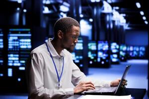 African american IT developer in data center using laptop to set up configuration management tools that enable automatic failover and load balancing, preventing server cabinets system crash photo