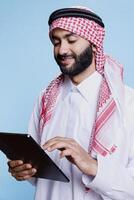 Smiling muslim person dressed in traditional clothes using digital tablet while engaging with social media messages. Cheerful arab person in islamic thobe and headdress using portable device in studio photo