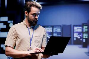 IT admin in hardware room containing server rackmounts, storage devices and networking units providing vast amounts of computing resources. Supervisor doing software maintenance using laptop photo