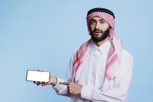 Muslim man holding smartphone with white screen in horizontal mode and pointing with finger. Arab person in traditional clothes showing empty touchscreen and looking at camera photo