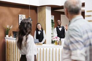 Caucasian elderly couple entering hotel entrance and approaching front desk for reservation assistance from multiethnic employees. Smiling receptionists warmly welcoming retired senior adults. photo