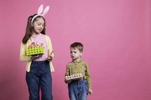 Sweet kids presenting their colorful easter ornaments on camera, little girl showing peace sign and posing next to her preschooler brother. Young siblings feeling happy about festive celebration. photo