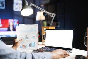 African american analyst examines white screen and report files , working remotely from home and using isolated copyspace display on portable computer. Expert looks at blank template and papers. photo