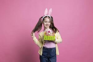 sonriente joven niño participación su Pascua de Resurrección preparativos con vistoso huevos como festivo primavera decoración, posando terminado rosado fondo. joven alegre niña vistiendo conejito orejas participación rosado mullido adornos foto