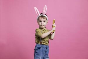 Happy smiling young kid playing with a plastic toy in studio, feeling joyful while he wears lovely bunny ears. Cheerful adorable boy standing against pink background, childhood innocence. photo