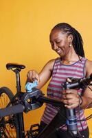 African american female cyclist inspects her broken bicycle frame on repair stand against isolated yellow backdrop. Black woman preparing to make adjustments and repairs with specialized equipment. photo