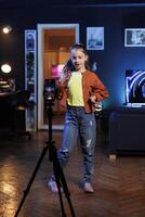 niña bailando y canto en personal estudio mientras disparo vídeo con Teléfono móvil en trípode para social medios de comunicación. alegre niño entretiene suscriptores con profesional coreografía grabado con móvil teléfono foto